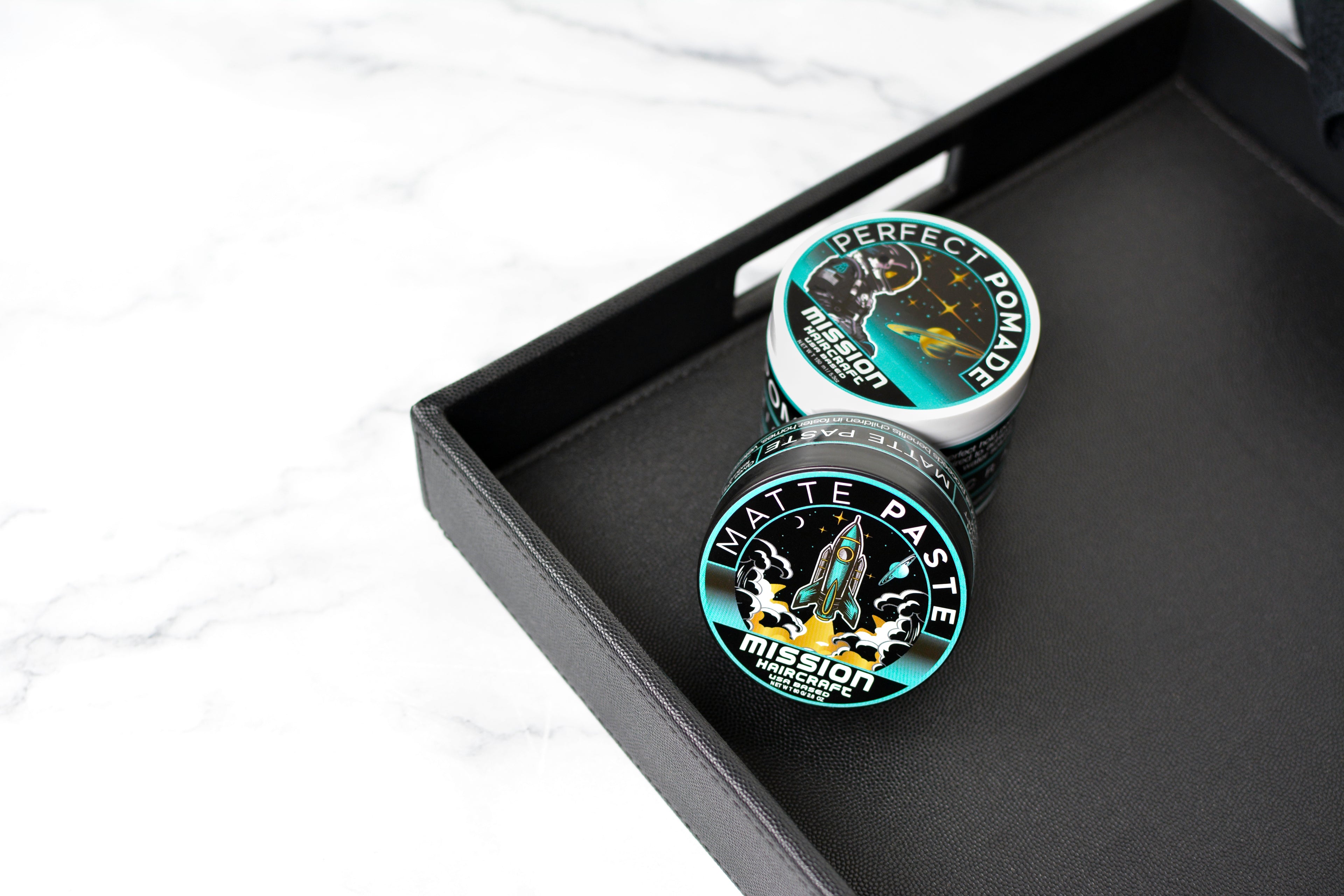 Two containers of 'Perfect Pomade' and 'Matte Paste Mission' on a black tray with a white background.